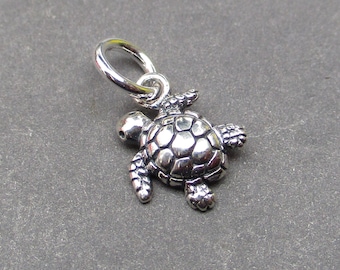 May include: A small, detailed silver turtle charm with a loop for attaching to a necklace or bracelet. The turtle has a textured shell and visible flippers and head. The charm is displayed against a gray background.
