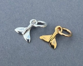 Può includere: Due piccoli ciondoli in metallo a forma di coda di balena, uno in tonalità argento lucido e l'altro in tonalità oro lucido. Ogni ciondolo presenta una superficie liscia e curva e include un piccolo anellino circolare fissato in alto per appenderlo.