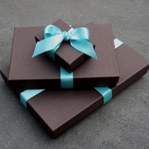 May include: Two rectangular dark brown gift boxes stacked on top of each other. The smaller top box is centred and features a bright light blue satin ribbon tied into a decorative bow. The larger bottom box is also wrapped with a matching blue ribbon.
