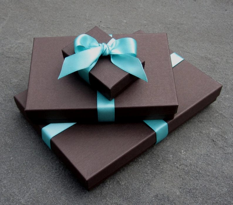 May include: Two rectangular dark brown gift boxes stacked on top of each other. The smaller top box is centred and features a bright light blue satin ribbon tied into a decorative bow. The larger bottom box is also wrapped with a matching blue ribbon.
