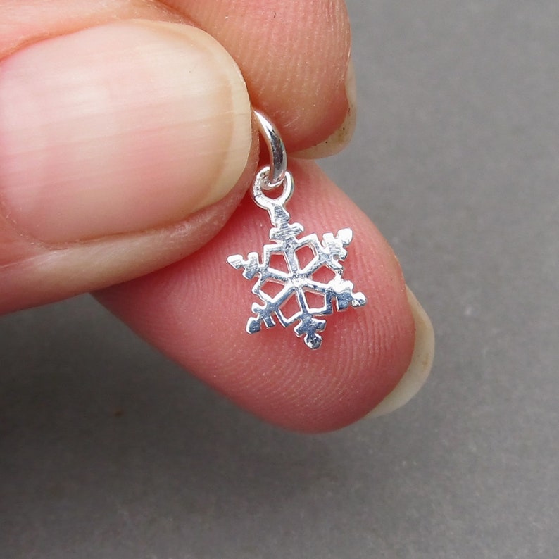 K&ouml;nnte beinhalten: Ein kleiner, silberfarbener Metall-Schneeflockenanh&auml;nger mit einem sechsstrahligen geometrischen Design, komplizierten Ausschnitten und einem runden Biegering an der Oberseite zum Aufh&auml;ngen. Der Anh&auml;nger hat eine polierte, reflektierende Oberfl&auml;che.