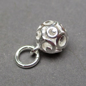 May include: Silver charm with a round, openwork design. The charm has a small loop for attaching to a bracelet or necklace.