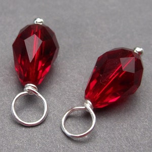 May include: Two red crystal teardrop shaped beads with silver wire loops. The beads are faceted and have a shiny finish.