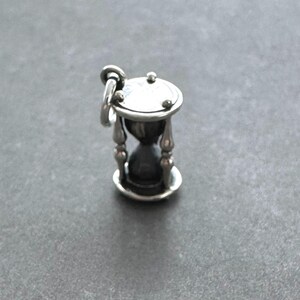 May include: A silver hourglass charm with a detailed design. The hourglass is filled with sand and has a small loop at the top for attaching to a necklace or bracelet.