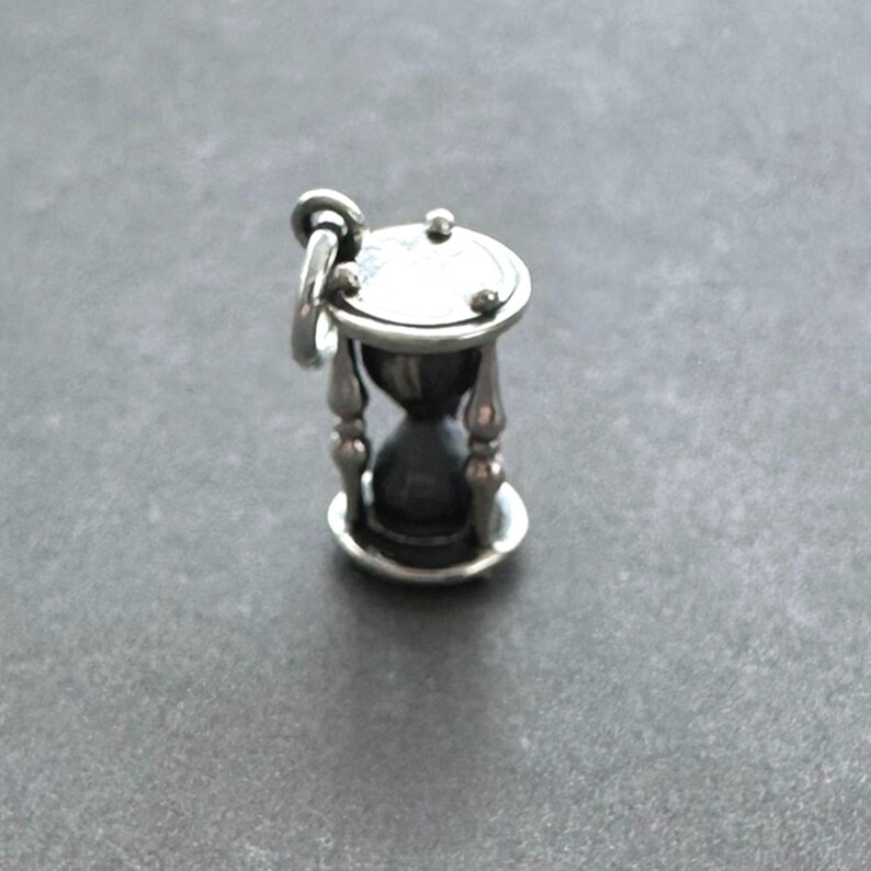 May include: A silver hourglass charm with a detailed design. The hourglass is filled with sand and has a small loop at the top for attaching to a necklace or bracelet.