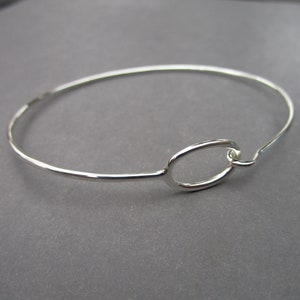 May include: A silver-colored bangle bracelet with an oval-shaped centerpiece. The bracelet has a simple, elegant design with a hook closure. The metal has a shiny finish, reflecting light. The bracelet is a piece of jewelry.