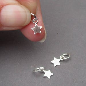 May include: Silver star charms with small hoops. One charm is held by a finger, while two others lay on a gray surface. The charms are small, with a simple star shape, and are likely for jewelry making.