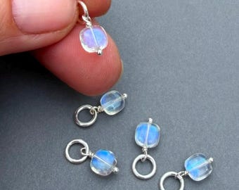 Puede incluir: Un conjunto de cuatro pequeños dijes, cada uno con una cuenta cuadrada, translúcida e iridiscente con un brillo azul, unida a una argolla de metal plateado. Las cuentas tienen un acabado liso y están conectadas por un alambre.