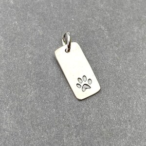 Pode incluir: Berloque de prata com um design de pegada de pata. O berloque &eacute; retangular e tem um la&ccedil;o na parte superior para prender a um colar ou pulseira.