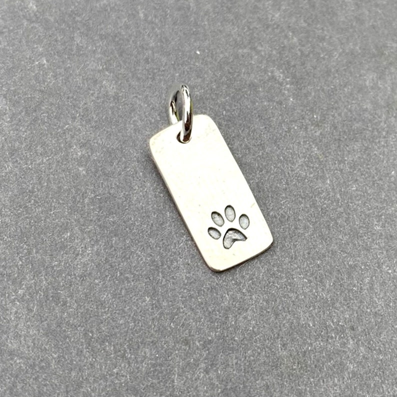Pode incluir: Berloque de prata com um design de pegada de pata. O berloque &eacute; retangular e tem um la&ccedil;o na parte superior para prender a um colar ou pulseira.