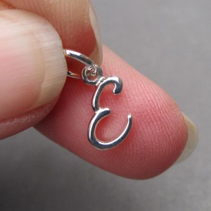 May include: Silver charm in the shape of the letter E. The charm has a loop at the top for attaching to a bracelet or necklace.