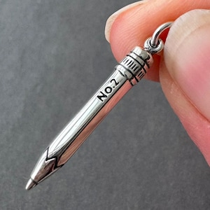 Op de afbeelding: Een zilverkleurige potloodbedel met de tekst "No.2" gegraveerd op de zijkant. De potloodbedel heeft een gedetailleerd gumuiteinde en een lus om aan een ketting of armband te bevestigen.