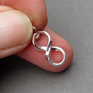 Könnte beinhalten: Ein kleiner, silberfarbener Metallanhänger in Form eines Unendlichkeitssymbols. Der Anhänger besteht aus einer durchgehenden, gedrehten Schleife mit einer glatten, reflektierenden Oberfläche und einem kleinen runden Ring an der Oberseite.