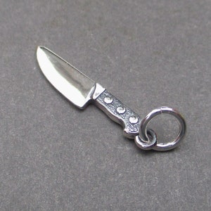 May include: A silver charm in the shape of a knife with a detailed handle and a ring for attaching to a bracelet or necklace.