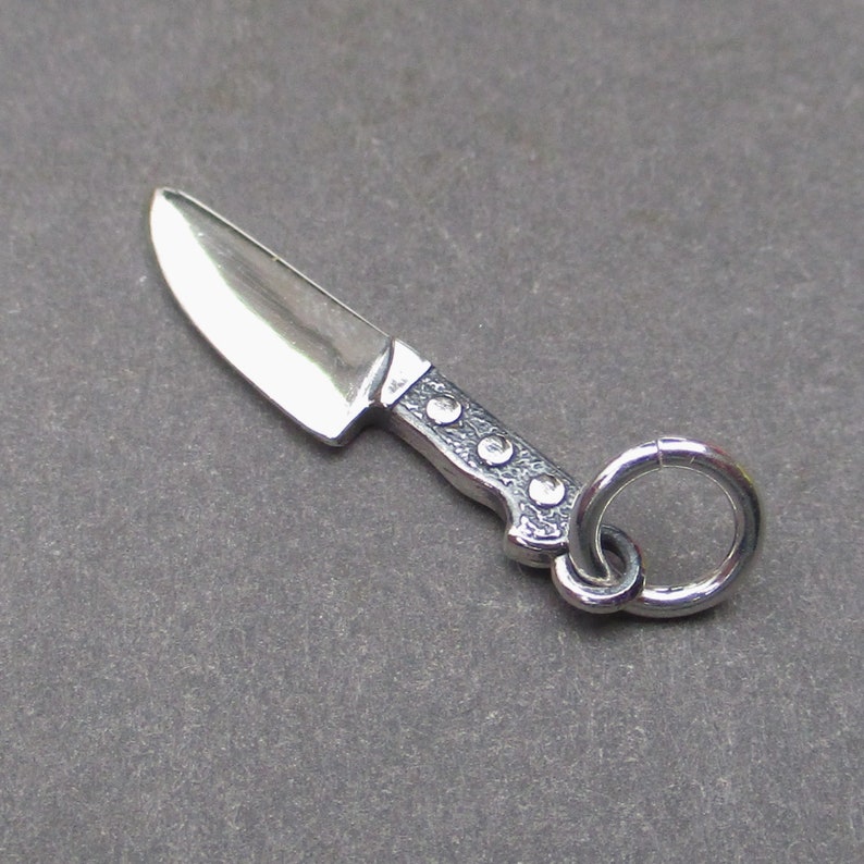 May include: A silver charm in the shape of a knife with a detailed handle and a ring for attaching to a bracelet or necklace.