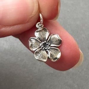 May include: A small, five-petaled silver flower charm featuring detailed, etched lines on each petal and a textured, beaded center. The pendant includes a small silver jump ring at the top for attachment to a necklace or bracelet chain.