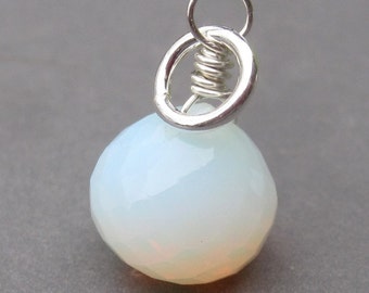 Puede incluir: Un colgante de piedra preciosa translúcida facetada, de color blanco opalescente con un sutil brillo iridiscente. La piedra está asegurada por un alambre de metal plateado que se conecta a una anilla circular plateada en la parte superior.