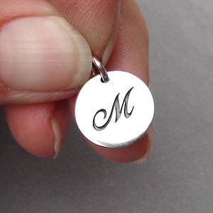 May include: Silver charm with a cursive letter M engraved on a round disc.