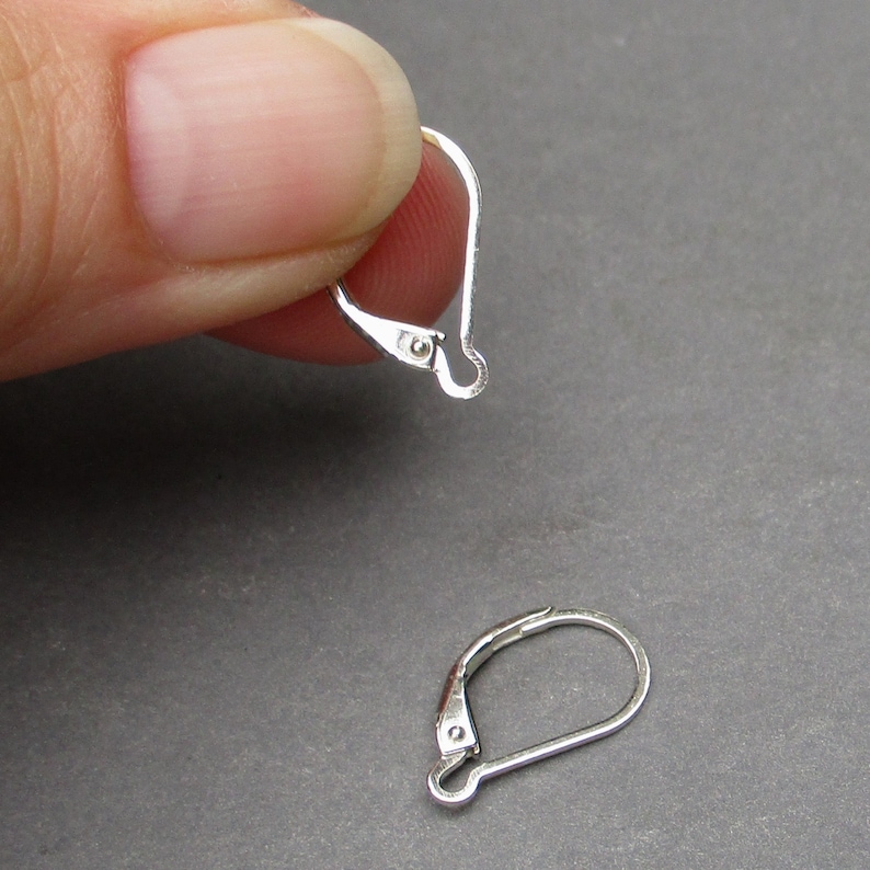 May include: Two silver-colored leverback earring findings. One is being held by a person's fingers. The findings are made of a shiny metal and have a simple, functional design. The background is a solid, neutral gray.