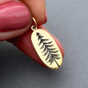May include: Gold-colored oval pendant with a black etched tree design. The pendant has a small loop at the top for a chain. The tree design is detailed and centered on the pendant. The pendant is held by a person.