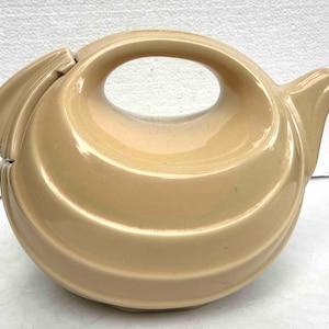 May include: A beige ceramic teapot with a rounded, horizontal body and a smooth, glossy finish. The design features a large, integrated handle at the top, a short spout on the right side, and a lid section on the left with a curved edge.