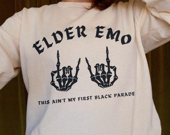 May include: Cream-colored sweatshirt with the text "ELDER EMO" in a gothic font, along with two black skeleton hand illustrations giving the middle finger. Below the text is the phrase "THIS AIN'T MY FIRST BLACK PARADE."