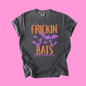 May include: A charcoal gray short-sleeve t-shirt featuring the orange text it's FRICKIN BATS, purple graphic illustrations of flying bats, and the purple text I love Halloween printed on the front of the garment.