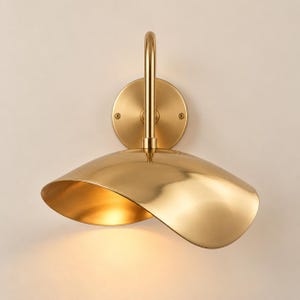 Könnte beinhalten: Eine Wandleuchte mit polierter Goldoberfläche. Sie besteht aus einer runden Wandplatte mit zwei sichtbaren Schrauben und einem geschwungenen, wellenförmigen Metallschirm, der das Licht nach unten lenkt. Das Design ist glatt, metallisch und symmetrisch.