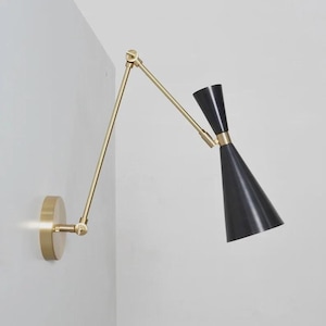 Könnte beinhalten: Eine schwarze und messingfarbene Wandleuchte mit verstellbarem Arm. Die Lampe hat einen konischen Schirm und einen kreisförmigen Sockel. Der Arm und der Sockel sind messingfarben, während der Schirm schwarz ist. Die Lampe ist an einer weißen Wand befestigt.