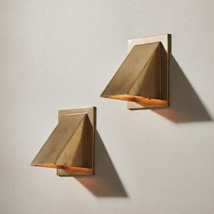 Può includere: Due applique da parete con paralume geometrico triangolare in ottone fissato a una piastra rettangolare. La finitura metallica ha una calda tonalità dorata e il design angolato dirige la luce verso il basso, creando ombre sulla parete.