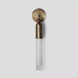 Puede incluir: Una lámpara de pared con una placa trasera circular de latón y una pantalla cilíndrica de vidrio esmerilado. La lámpara tiene un acabado de latón y un diseño minimalista, adecuado para la iluminación interior. La altura total es de unos 30 cm.