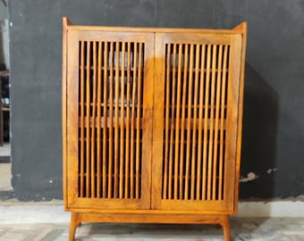 May include: A wooden cabinet with two doors featuring vertical slat detailing. The cabinet is a warm, medium brown color and stands on four short, angled legs. The doors have a slight arch at the top.