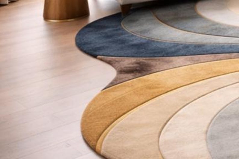 May include: A colorful, abstract area rug with a unique, flowing design. The rug features curved bands of color, including shades of blue, brown, beige, and gold. The rug's shape is irregular, adding to its artistic appeal. The rug is placed on a light-colored wooden floor.