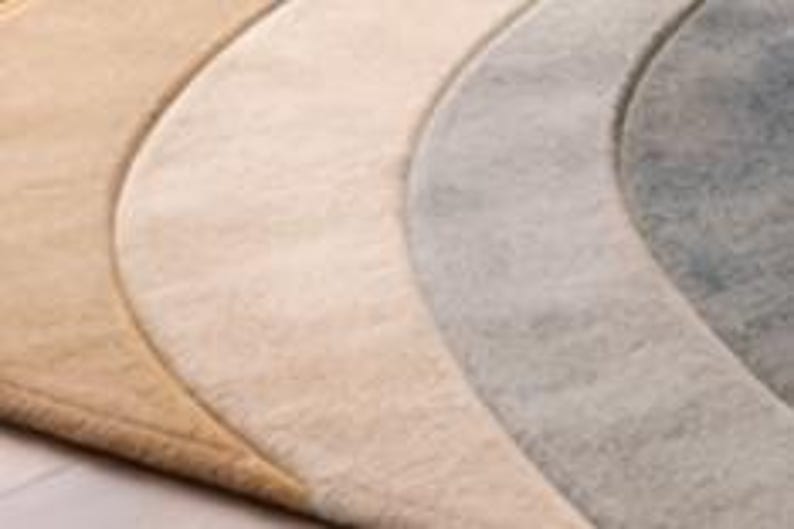 May include: A close-up of four crescent-shaped rugs in varying shades of beige, cream, gray, and blue. The rugs have a soft, textured appearance, with a subtle border outlining each shape. The arrangement suggests a layered or decorative display.