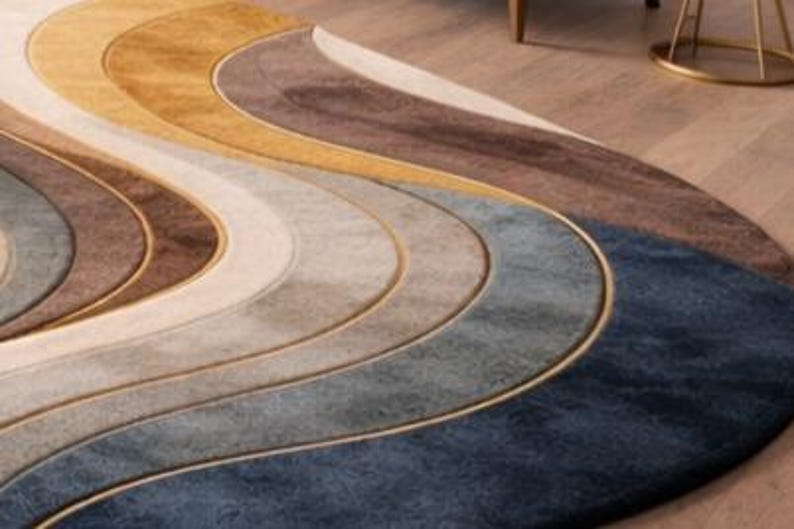 May include: A modern, abstract area rug with a wave-like design. The rug features a gradient of colors, including shades of blue, gray, brown, and gold. The rug is made of a soft material and has a gold border. The rug is a decorative home decor item.