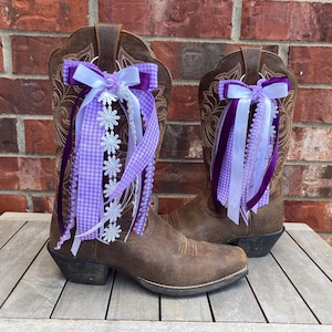 May include: A pair of brown leather cowboy boots with decorative purple and white ribbons and bows. The boots are displayed on a wooden surface against a brick background. The ribbons include gingham and lace details.