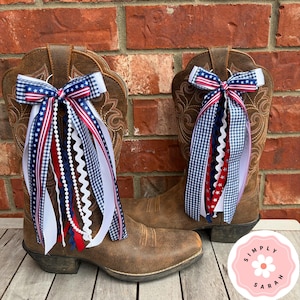 May include: A pair of brown leather cowboy boots adorned with decorative bows made of red, white, and blue ribbons. The ribbons feature patterns including stars, stripes, and gingham checks. A pink circular logo with the text Simply Sarah is visible.