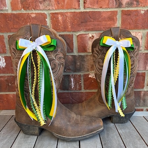 May include: A pair of brown leather cowboy boots with decorative stitching. Each boot is adorned with a bow made of white, yellow, and green ribbons, with matching streamers. The boots are set against a brick wall and wooden surface.
