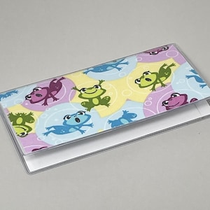 May include: A rectangular checkbook cover with a clear plastic exterior. The cover features a colorful pattern of cartoon frogs in various colors, including green, purple, and blue, set against a pastel background.