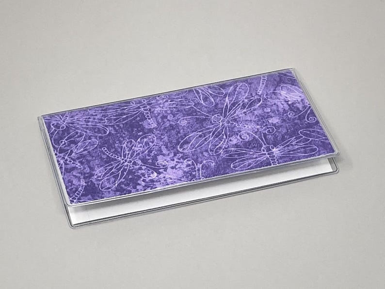 May include: A purple checkbook cover with a dragonfly pattern. The cover has a clear plastic frame and a white interior. The dragonfly design is in white, with a textured purple background.