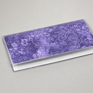 May include: A checkbook cover with a purple background and white dragonfly design. The cover is made of a textured material and has a clear plastic overlay. The checkbook is open, revealing the white check paper.