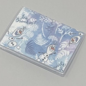 Puede incluir: Un porta tarjetas de plástico transparente con un patrón azul y blanco con Olaf de la película Frozen. El patrón incluye el texto "Ice Breaker!" y diseños florales blancos.