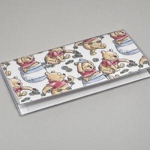 May include: A clear plastic checkbook cover featuring a repeating pattern of Winnie the Pooh characters. The illustrations show Pooh Bear in various poses, wearing a red shirt, with daisies on a white background.