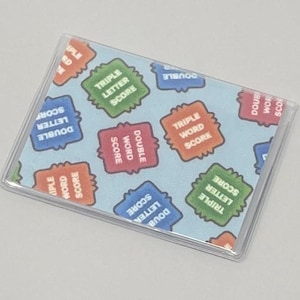 Può includere: Una custodia in plastica trasparente con sfondo blu e un motivo di quadrati colorati. I quadrati hanno il testo "Double Letter Score", "Triple Letter Score", "Double Word Score" e "Triple Word Score".