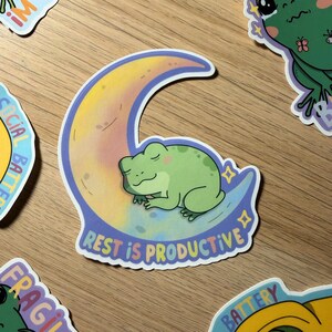 Op de afbeelding: Een sticker met een lichtgroene kikker die slaapt op een halve maan met een verloop van geel, roze en blauw. De tekst "REST IS PRODUCTIVE" staat eronder. Andere kikkerstickers zijn zichtbaar.