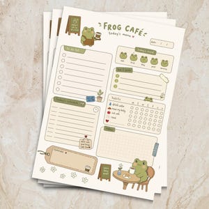 May include: A cream-coloured daily planner sheet titled Frog Café today's menu, featuring green frog illustrations, a to-do list, a schedule, habit trackers, and goal sections. The layout includes small icons, a date field, and notes area.