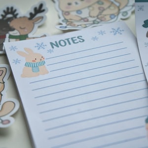 Könnte beinhalten: Notizblock mit dem Wort „NOTES“ in Hellblau, mit einem Cartoon-Hasen mit einem blau-weiß gestreiften Schal. Der Notizblock hat blaue horizontale Linien und Schneeflocken-Illustrationen. Tieraufkleber im Hintergrund.