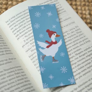 Könnte beinhalten: Blauer Lesezeichen mit einer weißen Gans mit roter Mütze und Schal, die Schlittschuh läuft. Das Lesezeichen hat weiße Schneeflocken und liegt auf einem offenen Buch.