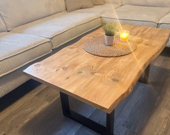 May include: A natural wood coffee table with a live edge design. The table has a light brown finish and sits on black metal legs. A small potted plant and a lit candle rest on a woven placemat on the table's surface.
