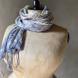 Op de afbeelding: Blauw en crèmekleurige sjaal met een paisley- en bloemenpatroon, gedrapeerd over een mannequin. De sjaal heeft een gefranjerde rand en is gemaakt van een lichte stof. Een modeaccessoire.